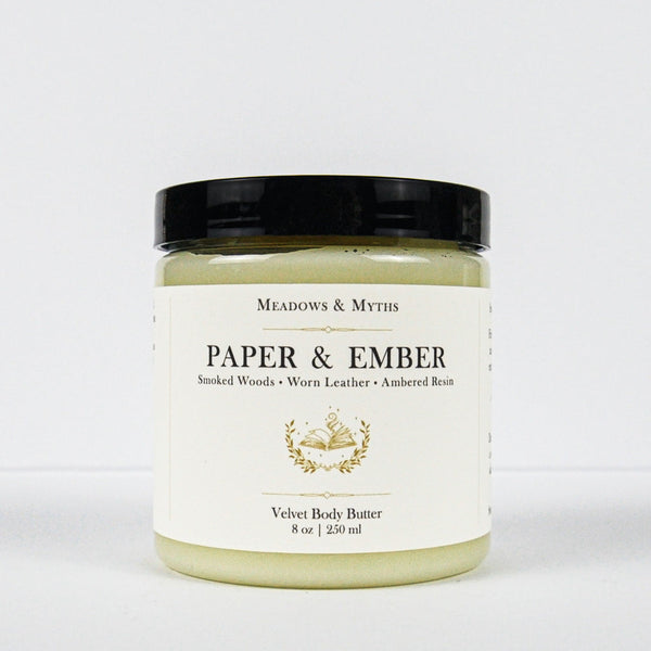 A jar of Meadows & Myth's 'Paper & Ember' velvet body butter, featuring a cream-colored label with gold detailing and a black lid, set against a plain white background.
