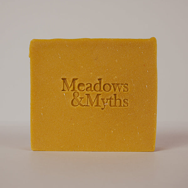 A rectangular bar of mustard yellow soap with the brand name 'Meadows & Myths' embossed in the center, photographed against a neutral light gray background.