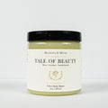 A jar of Meadows & Myth's 'Tale of Beauty' velvet body butter, featuring a light olive green body butter visible through the clear jar, a cream-colored label with gold detailing, and a black lid, set against a plain white background.
