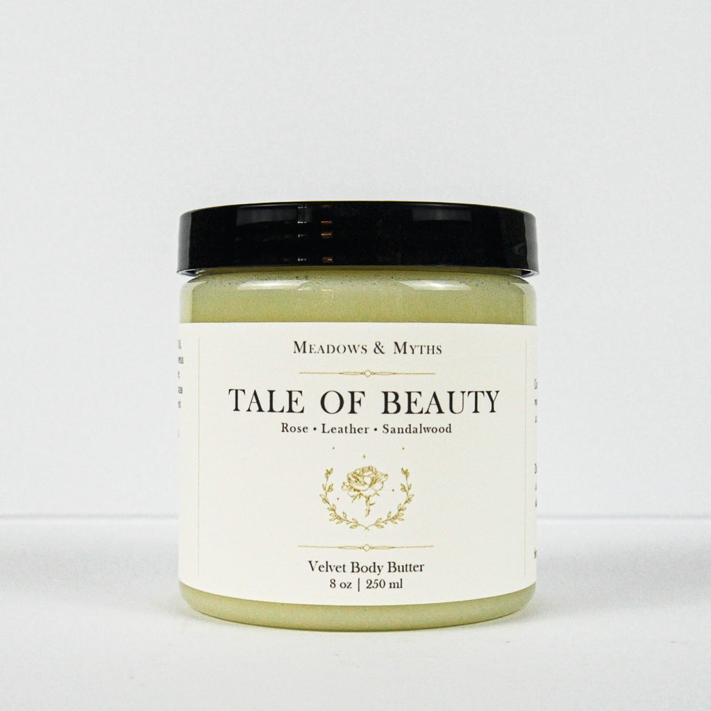 A jar of Meadows & Myth's 'Tale of Beauty' velvet body butter, featuring a light olive green body butter visible through the clear jar, a cream-colored label with gold detailing, and a black lid, set against a plain white background.