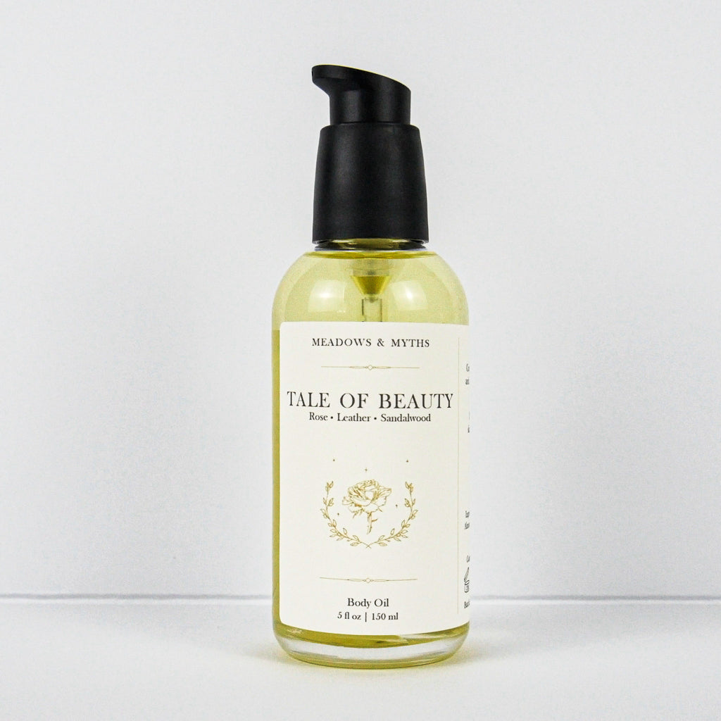 A bottle of Meadows & Myth's 'Tale of Beauty' rose, leather, and sandalwood body oil. The clear bottle shows the pale yellow oil inside, with a cream-colored label and a black pump dispenser, set against a plain white background.