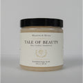 A jar of Meadows & Myth's 'Tale of Beauty' emulsified sugar scrub, featuring a cream-colored label with gold detailing and a black lid, set against a neutral light gray background.