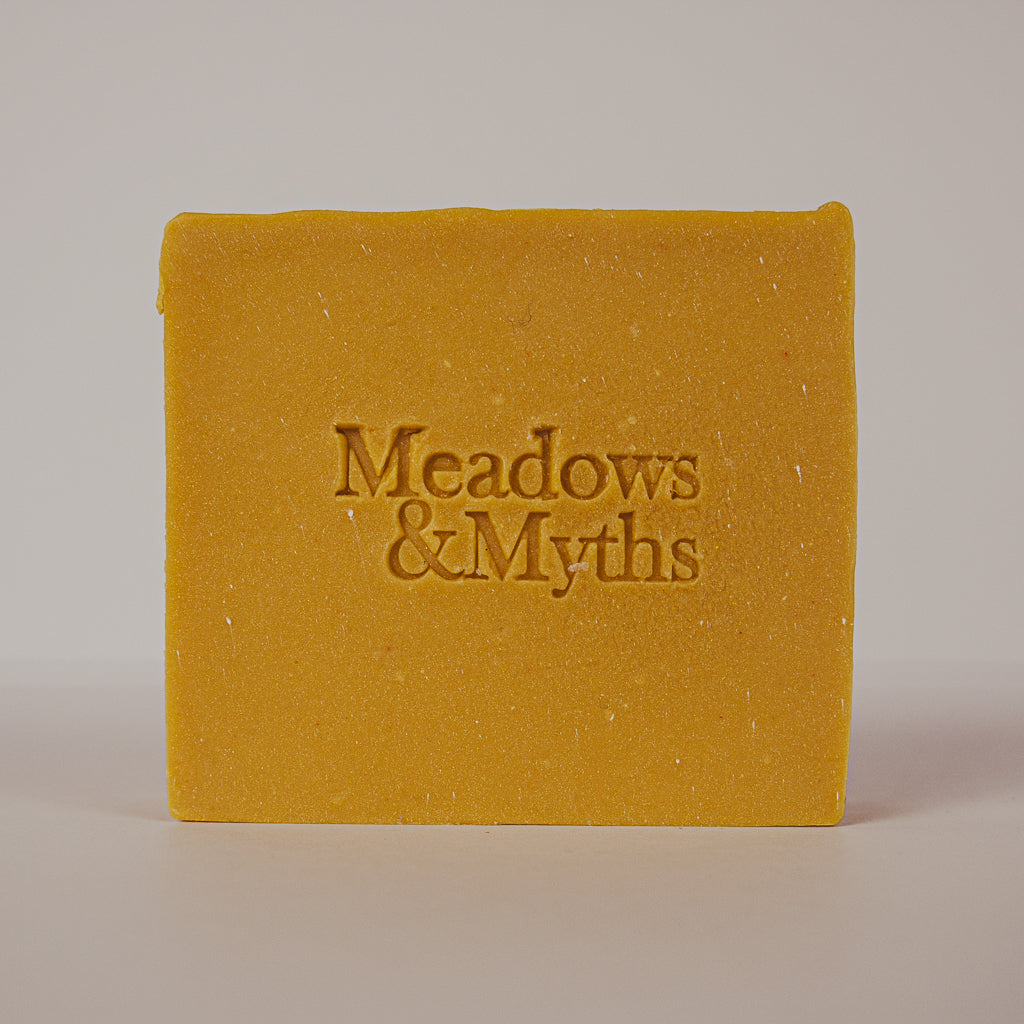 A rectangular bar of mustard yellow soap with the brand name 'Meadows & Myths' embossed in the center, photographed against a neutral light gray background.