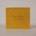 A rectangular bar of mustard yellow soap with the brand name 'Meadows & Myths' embossed in the center, photographed against a neutral light gray background.