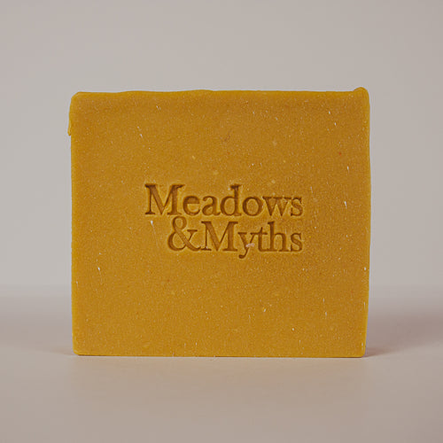 A rectangular bar of mustard yellow soap with the brand name 'Meadows & Myths' embossed in the center, photographed against a neutral light gray background.