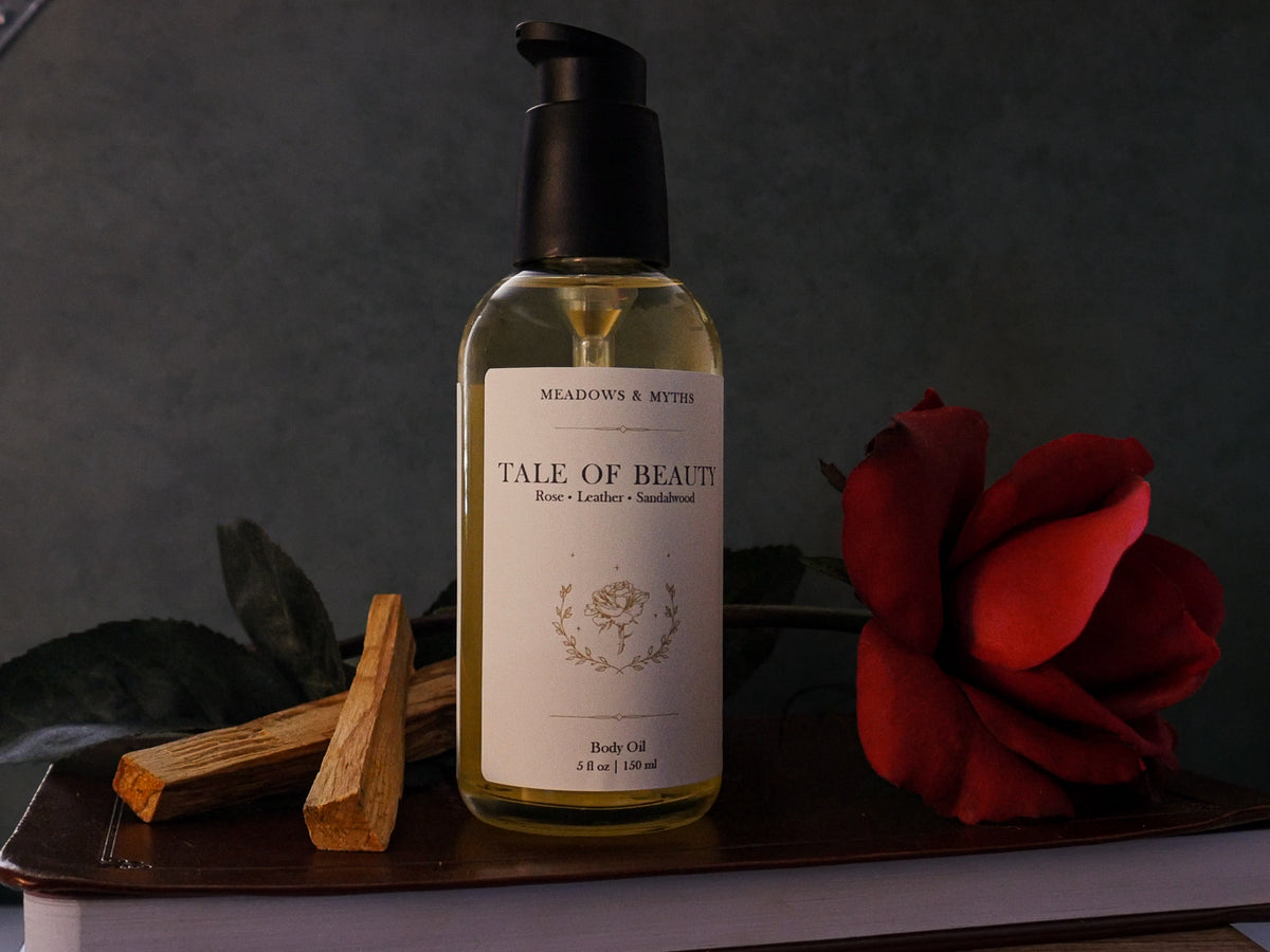 A glass bottle with a black pump of Meadows & Myths 'Tale of Beauty' body oil, styled with a red rose and sandalwood sitting on top of a leather book, against a dark background.