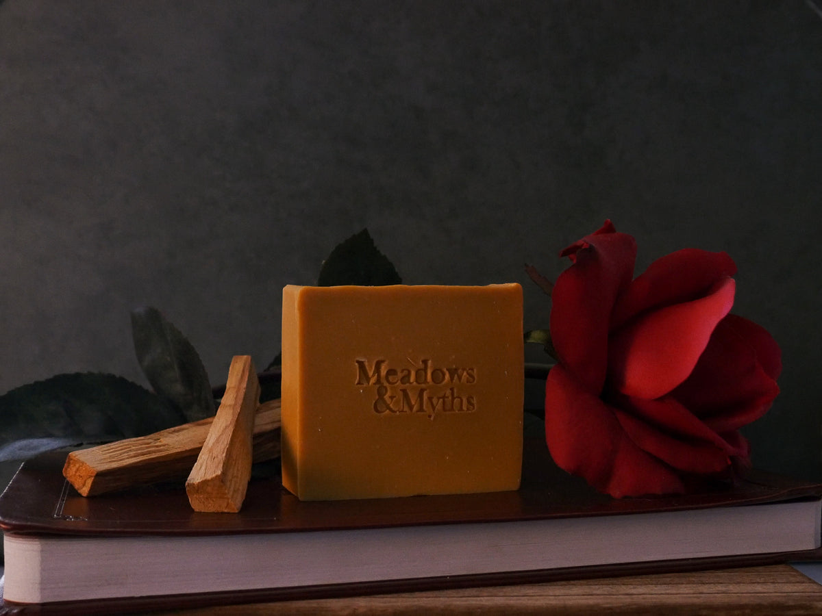 A rectangular bar of mustard-yellow soap with 'Meadows & Myths' embossed on the front, styled with a red rose and sandalwood sitting on top of a leather book, against a dark background.