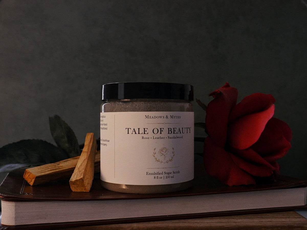 A jar of Meadows & Myths 'Tale of Beauty' emulsified sugar scrub , styled with a red rose and sandalwood sitting on top of a leather book, against a dark background.