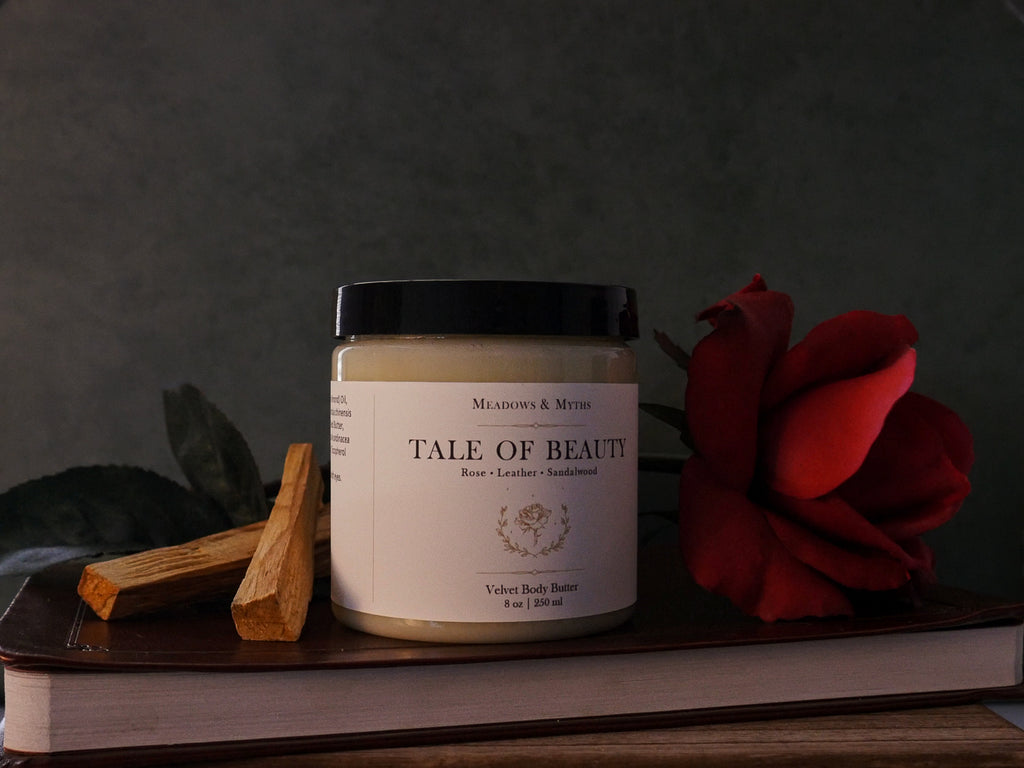 A jar of Meadows & Myths Velvet Body Butter "Tale of Beauty, styled with a red rose and sandalwood sitting on top of a leather book, against a dark background.