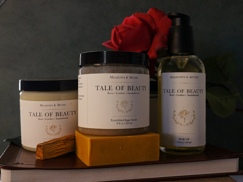 A collection of Meadows & Myths 'Tale of Beauty' bath and body products, including a yellow bar of soap, a pump bottle of Body Oil, two jars: Body Butter and Sugar Scrub,, styled with a red rose and sandalwood sitting on top of a leather book, against a dark background.