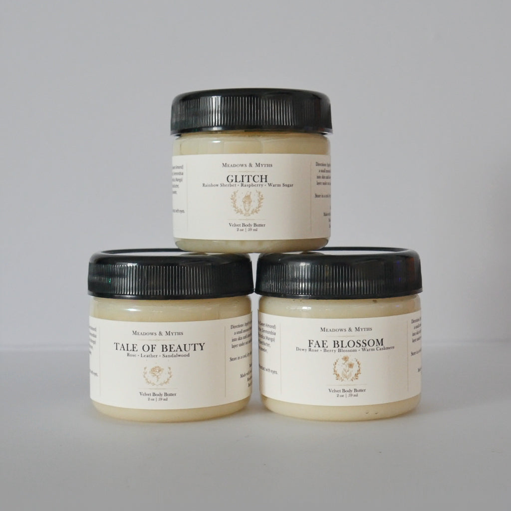 Three jars of Meadows & Myths body butter products stacked in a pyramid on a light gray background. The top jar is 'Glitch' and the bottom two are 'Tale of Beauty' and 'Fae Blossom'.