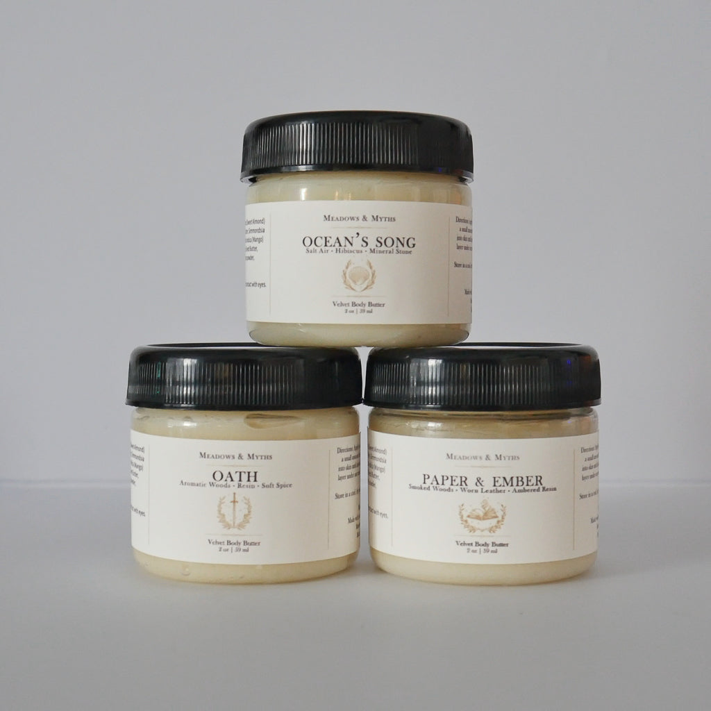 Three jars of Meadows & Myths body butter products stacked in a pyramid on a light gray background. The top jar is 'Ocean's Song' and the bottom two are 'Oath' and 'Paper & Ember'.