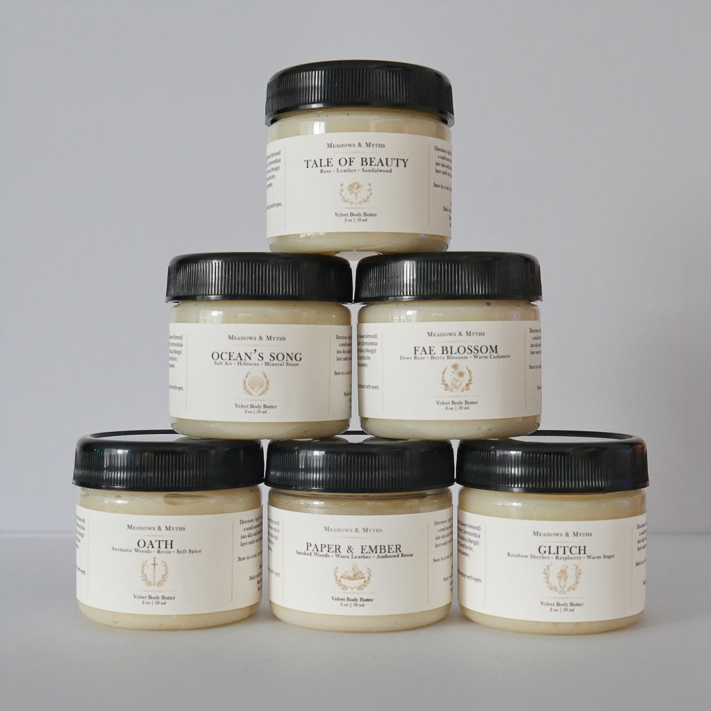 Six jars of Meadows & Myths body butter arranged same jars in a pyramid stack on a light gray background. The jars have a  cream-colored label with black lids and feature labels for Tale of Beauty, Ocean's Song, Fae Blossom, Oath, Paper & Ember, and Glitch.