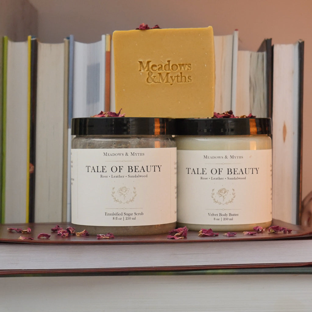 Meadows & Myths Tale of Beauty body care set featuring a jar of body butter, jar of sugar scrub, yellow rectangle soap. Styled on a stack of books with dried rose petals scattered around it and a row of books in the background.