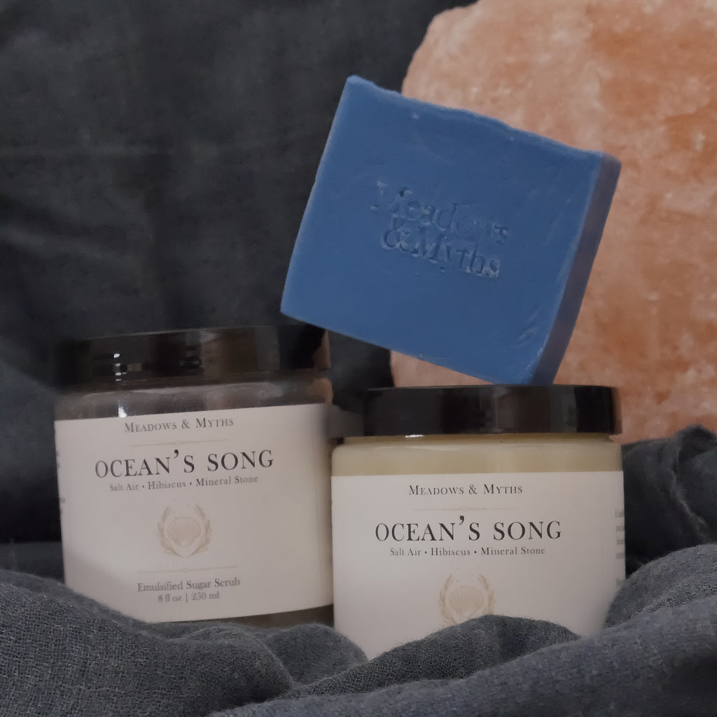 Meadows & Myths 'Ocean's Song' scented body care set featuring a blue rectangular soap bar, a jar of sugar scrub, and a jar of body butter, styled with a pink salt stone and dark gray-blue fabric.