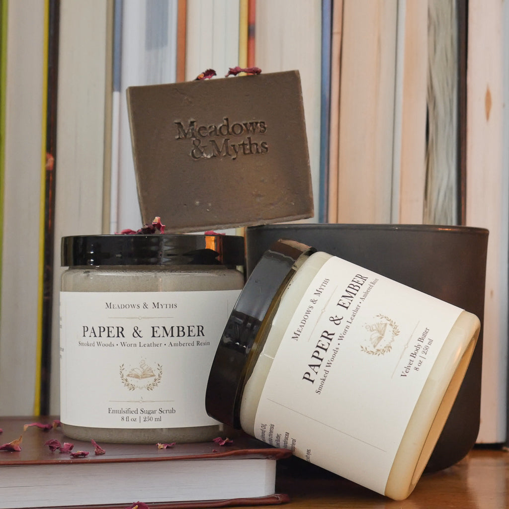 Meadows & Myths Paper & Ember body care set featuring a jar of body butter, jar of sugar scrub, brown rectangle soap. Styled on a wooden surface with dried rose petals scattered around it. They sit on top of a closed leather bound book, with a dark green candle and a row of books in the background.