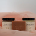 Meadows & Myths 'Fae Blossom' scented body care set featuring a terracotta rectangular soap bar with "Meadows & Myths" embossed on it, a jar of sugar scrub, and a jar of body butter, sitting on pink fabric with a white background.