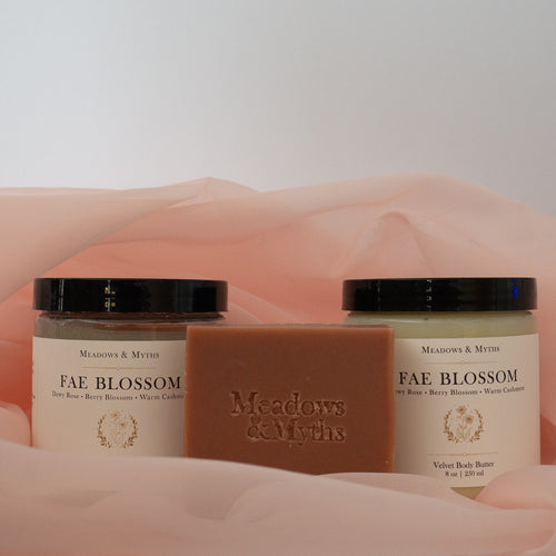 Meadows & Myths 'Fae Blossom' scented body care set featuring a terracotta rectangular soap bar with "Meadows & Myths" embossed on it, a jar of sugar scrub, and a jar of body butter, sitting on pink fabric with a white background.