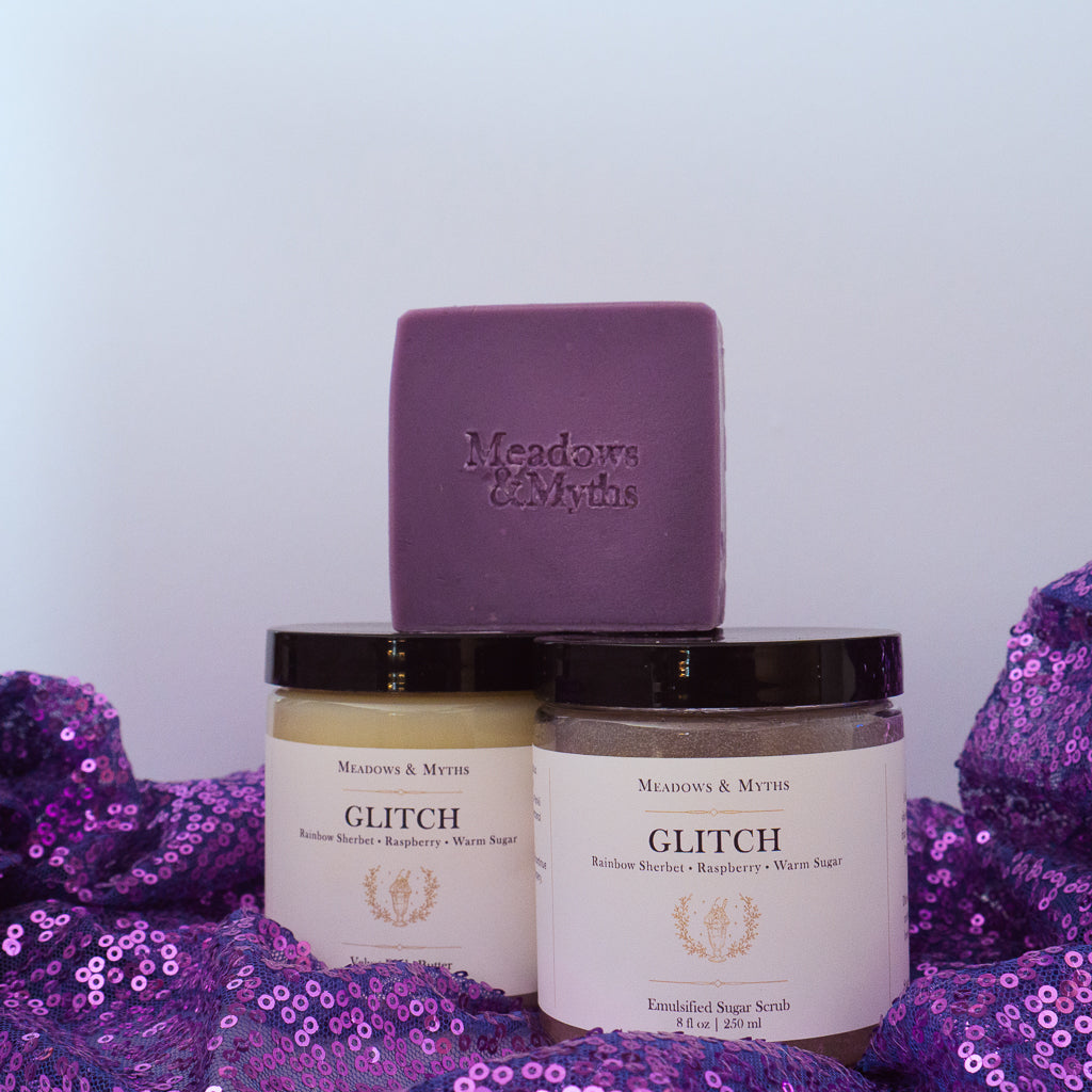 Meadows & Myths 'Glitch' scented body care set featuring a purple rectangular soap bar with "Meadows & Myths" embossed on it, a jar of sugar scrub, and a jar of body butter, sitting on purple sequins fabric.