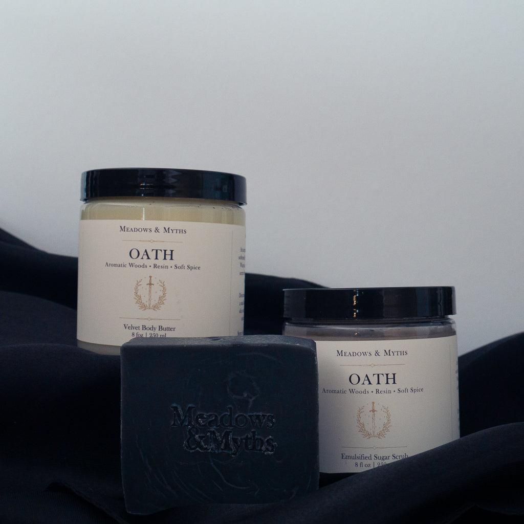 Meadows & Myths 'Oath' scented body care set featuring a black rectangular soap bar with "Meadows & Myths" embossed on it, a jar of sugar scrub, and a jar of body butter, sitting on black fabric with a white background with a white background.