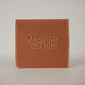 A square bar of terracotta-colored soap with the brand name 'Meadows & Myths' embossed on the front, set against a neutral light gray background.