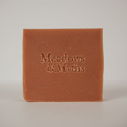 A square bar of terracotta-colored soap with the brand name 'Meadows & Myths' embossed on the front, set against a neutral light gray background.