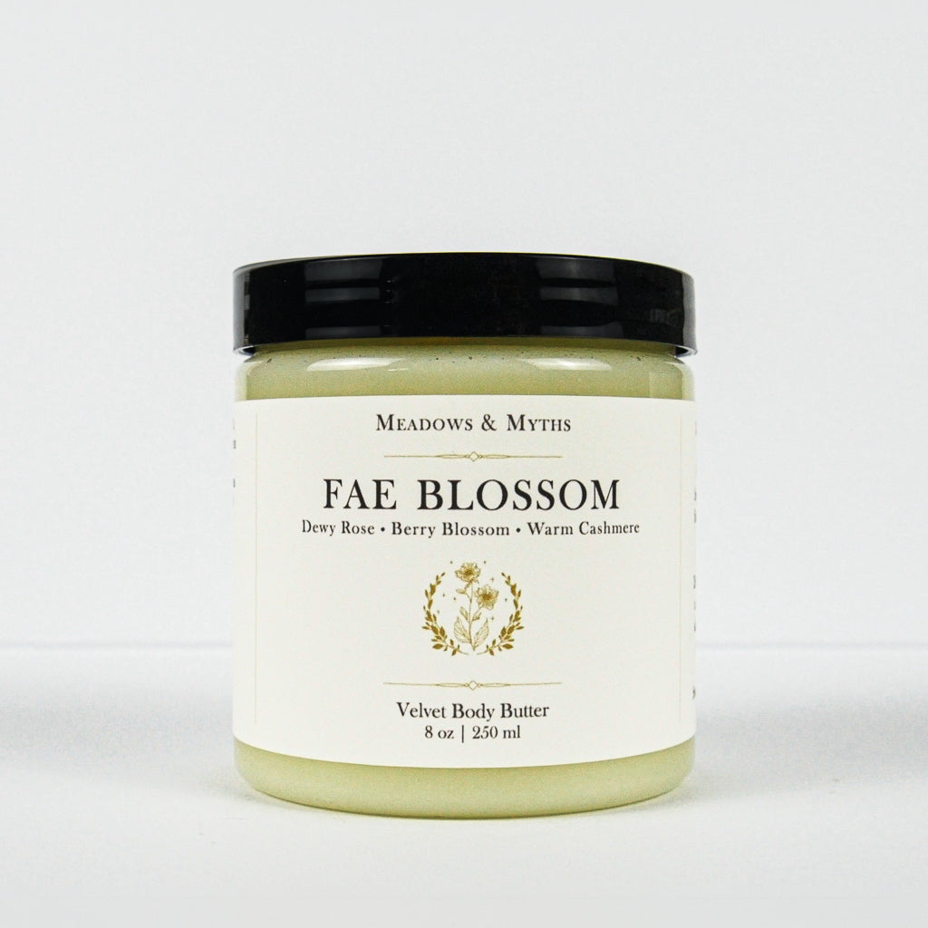 A jar of Meadows & Myth's 'Fae Blossom' velvet body butter, featuring a cream-colored label with gold detailing and a black lid, set against a plain white background.
