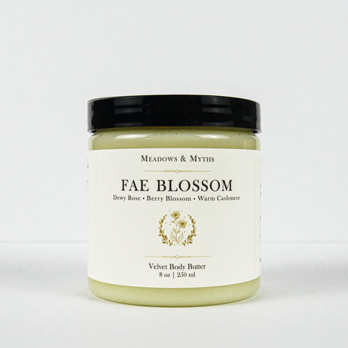 A jar of Meadows & Myth's 'Fae Blossom' velvet body butter, featuring a cream-colored label with gold detailing and a black lid, set against a plain white background.
