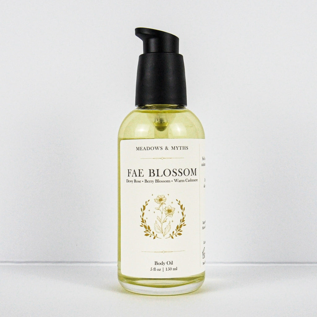 A bottle of Meadows & Myth's Fae Blossom body oil, featuring a clear glass bottle filled with pale yellow oil, a cream-colored label with gold botanical illustrations, and a black pump dispenser, set against a plain white background.