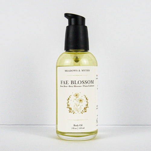 A bottle of Meadows & Myth's Fae Blossom body oil, featuring a clear glass bottle filled with pale yellow oil, a cream-colored label with gold botanical illustrations, and a black pump dispenser, set against a plain white background.