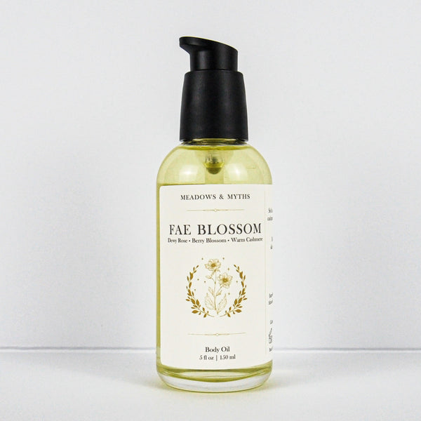 A bottle of Meadows & Myth's Fae Blossom body oil, featuring a clear glass bottle filled with pale yellow oil, a cream-colored label with gold botanical illustrations, and a black pump dispenser, set against a plain white background.