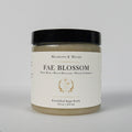 A jar of Meadows & Myth's 'Fae Blossom' emulsified sugar scrub, featuring a cream-colored label with gold detailing and a black lid, set against a plain light gray background.