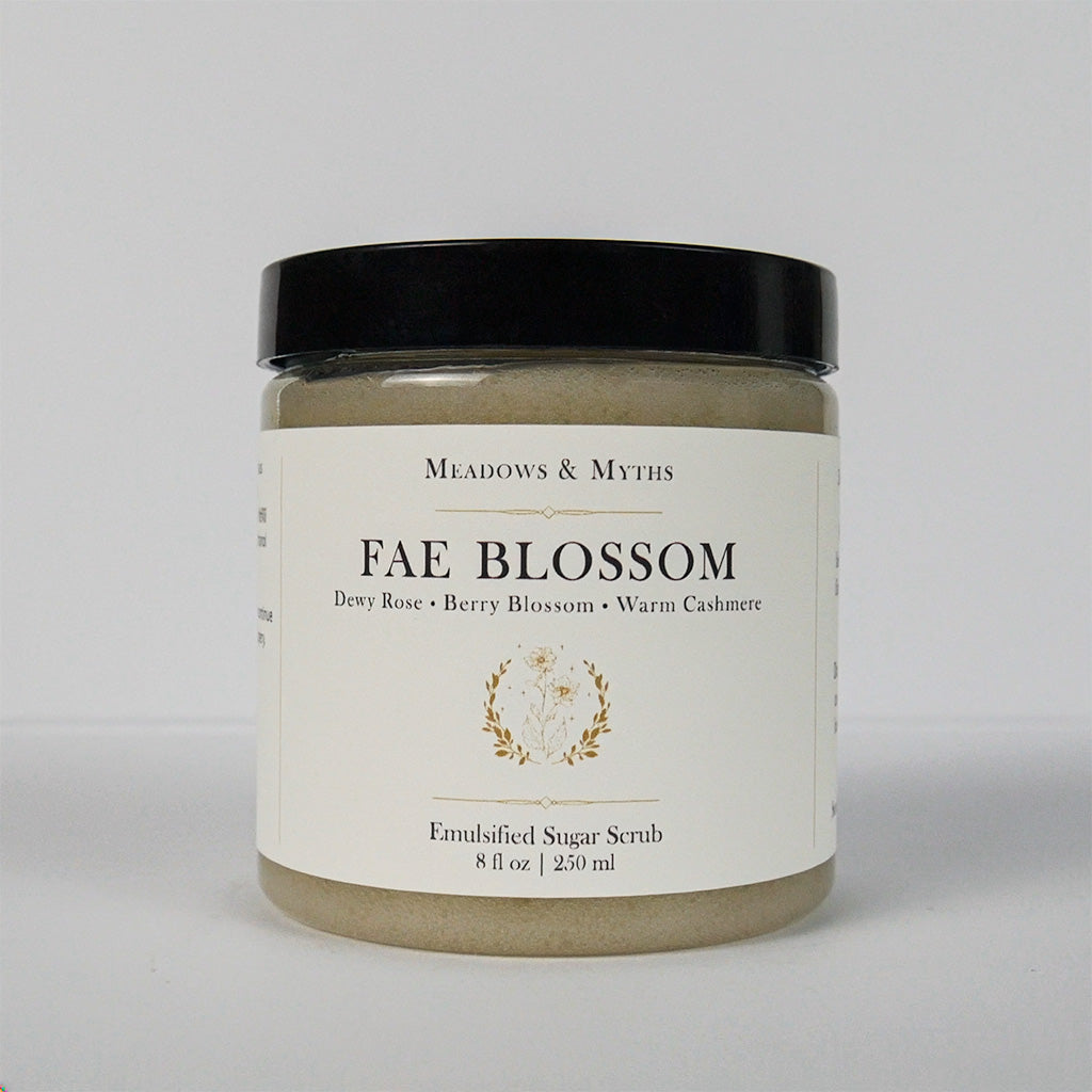 A jar of Meadows & Myth's 'Fae Blossom' emulsified sugar scrub, featuring a cream-colored label with gold detailing and a black lid, set against a plain light gray background.