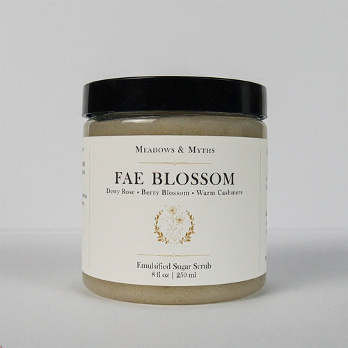 A jar of Meadows & Myth's 'Fae Blossom' emulsified sugar scrub, featuring a cream-colored label with gold detailing and a black lid, set against a plain light gray background.