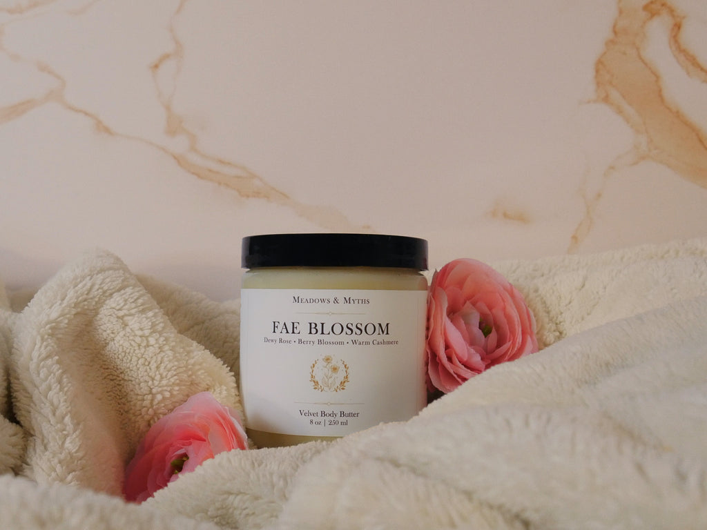 A jar of Meadows & Myths 'Fae Blossom' Velvet Body Butter , styled on a cream fleece blanket with pink flowers against a beige marble background.