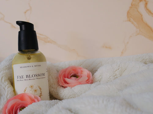 A glass bottle with a black pump of Meadows & Myths 'Fae Blossom' body oil, styled on a cream fleece blanket with pink flowers against a beige marble background.