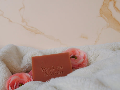 A rectangular bar of terracotta-colored soap with the brand name 'Meadows & Myths' embossed on the front, resting on a white plush towel surrounded by pink flowers against a beige marble-patterned background.