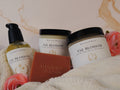 A jar of Meadows & Myths 'Fae Blossom' Velvet Body Butter, a jar of emulsified sugar scrub, a glass bottle with a black pump of body oil, a rectangular bar of terracotta-colored soap with the brand name 'Meadows & Myths' embossed on the front, styled on a cream fleece blanket with pink flowers against a beige marble background.