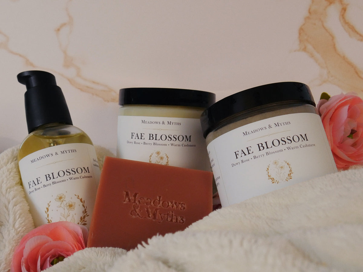 A jar of Meadows & Myths 'Fae Blossom' Velvet Body Butter, a jar of emulsified sugar scrub, a glass bottle with a black pump of body oil, a rectangular bar of terracotta-colored soap with the brand name 'Meadows & Myths' embossed on the front, styled on a cream fleece blanket with pink flowers against a beige marble background.