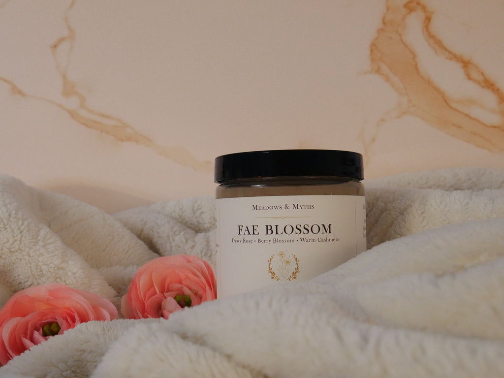 A jar of Meadows & Myths 'Fae Blossom' emulsified sugar scrub, styled on a cream fleece blanket with pink flowers against a beige marble background.