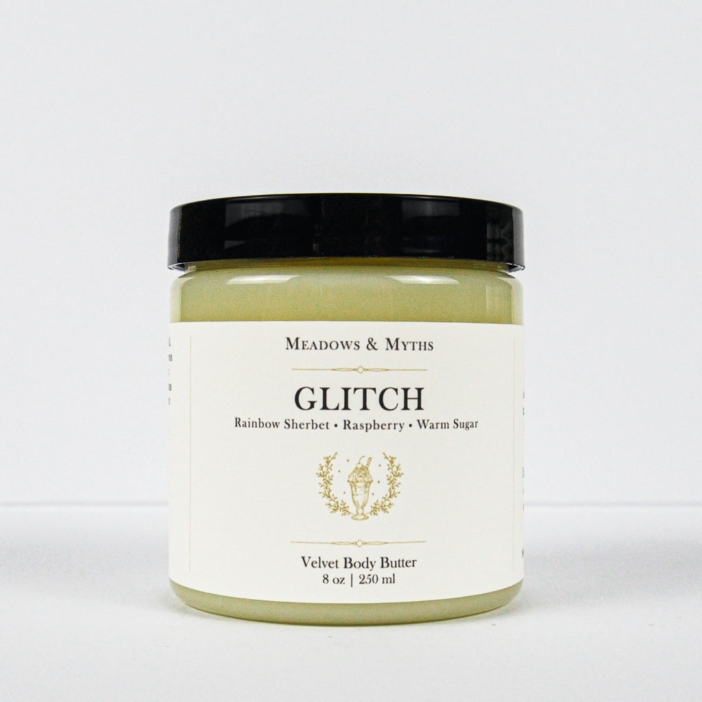 A jar of Meadows & Myth's Glitch Velvet Body Butter with a black lid and a cream-colored label, set against a plain white background.