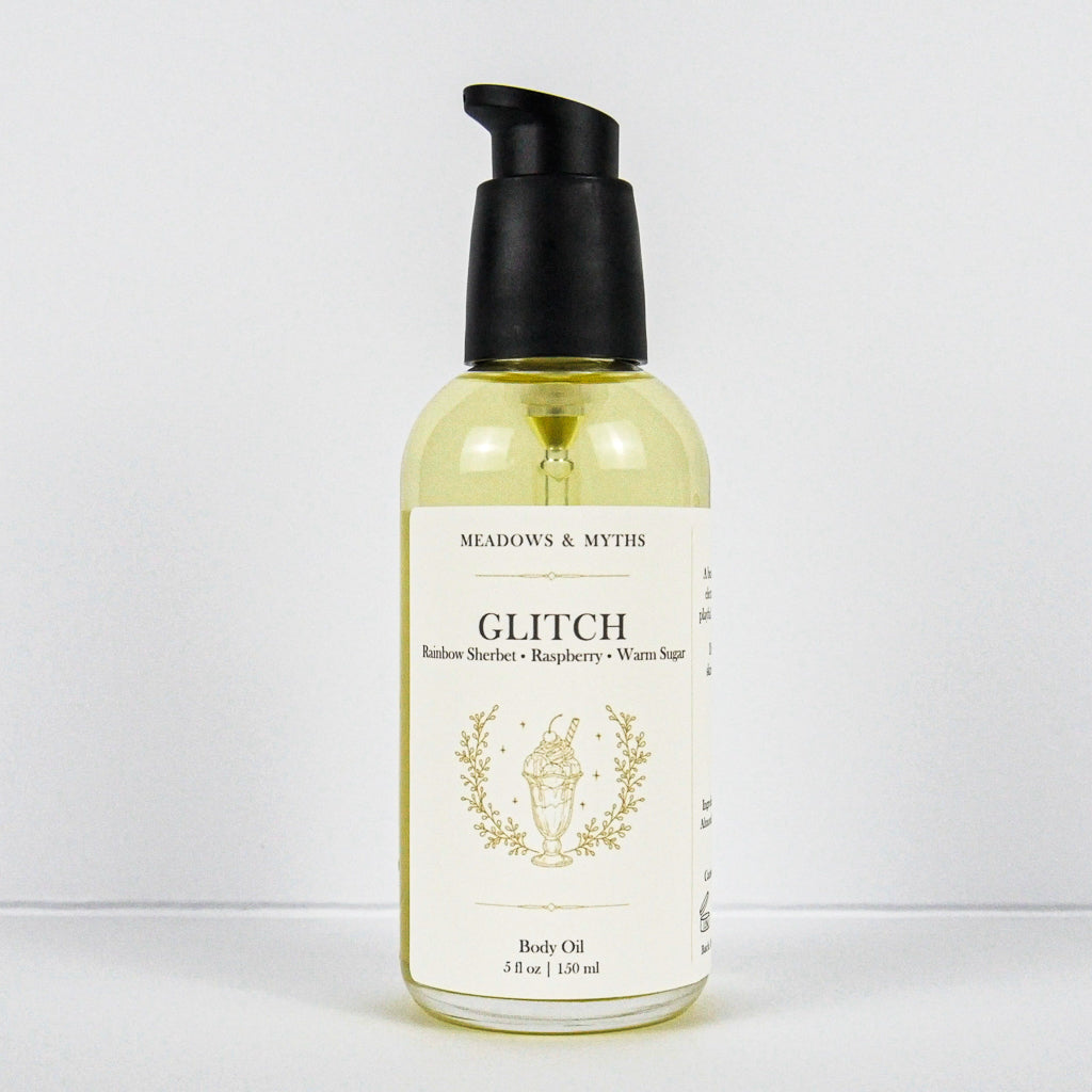 A bottle of Meadows & Myth's Glitch body oil, featuring a clear bottle filled with pale yellow oil, a black pump dispenser, and a cream-colored label with gold text and illustration, set against a plain white background.