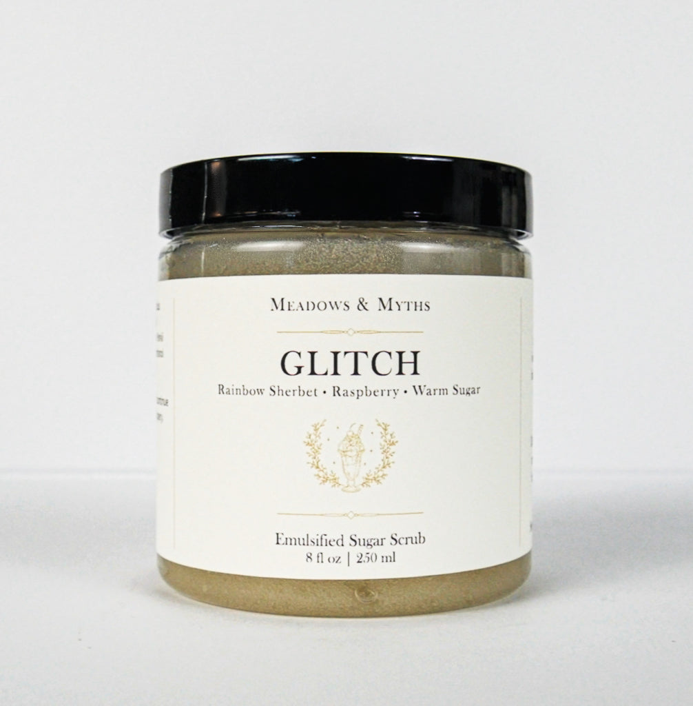 A jar of Meadows & Myth's Glitch Emulsified Sugar Scrub, featuring a white label with gold lettering and a black lid, set against a plain white background.