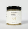 A jar of Meadows & Myth's Glitch Emulsified Sugar Scrub, featuring a white label with gold lettering and a black lid, set against a plain white background.