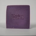 A single rectangular purple bar of soap with the brand name 'Meadows & Myths' embossed on the front, photographed against a neutral light gray background.