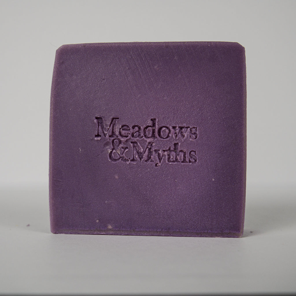 A single rectangular purple bar of soap with the brand name 'Meadows & Myths' embossed on the front, photographed against a neutral light gray background.