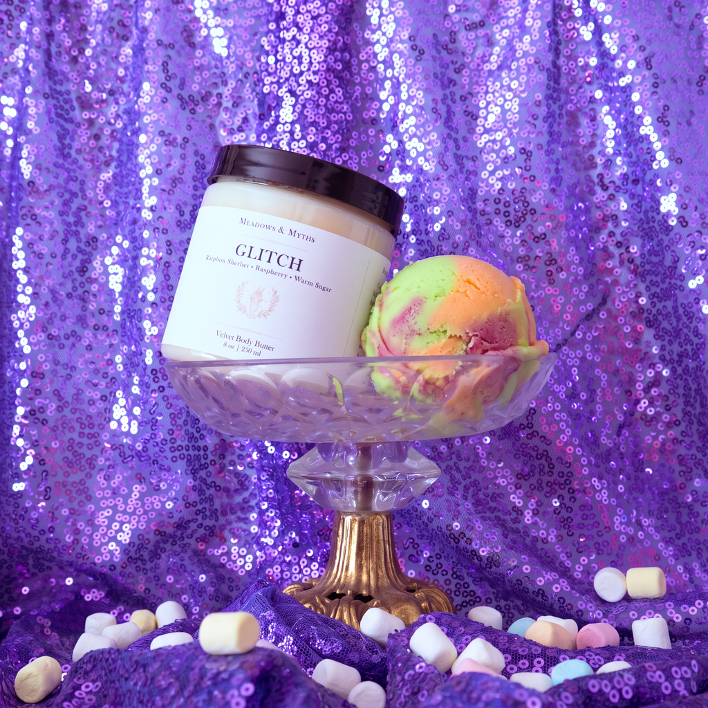 Meadows & Myths 'Glitch' jar of sugar scrub. Sitting on purple sequins fabric with the same fabric draped in the background. The body butter sits in a glass raised dish with a scoop of rainbow sherbet. Colorful marshmallows are scattered on the ground.  