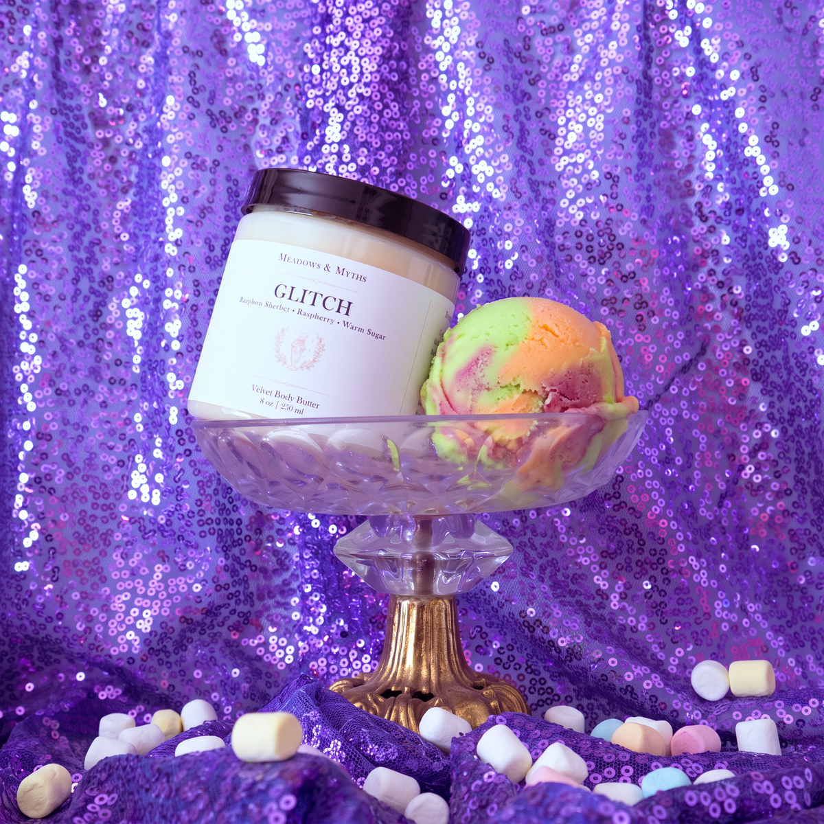 Meadows & Myths 'Glitch' jar of sugar scrub. Sitting on purple sequins fabric with the same fabric draped in the background. The body butter sits in a glass raised dish with a scoop of rainbow sherbet. Colorful marshmallows are scattered on the ground.  