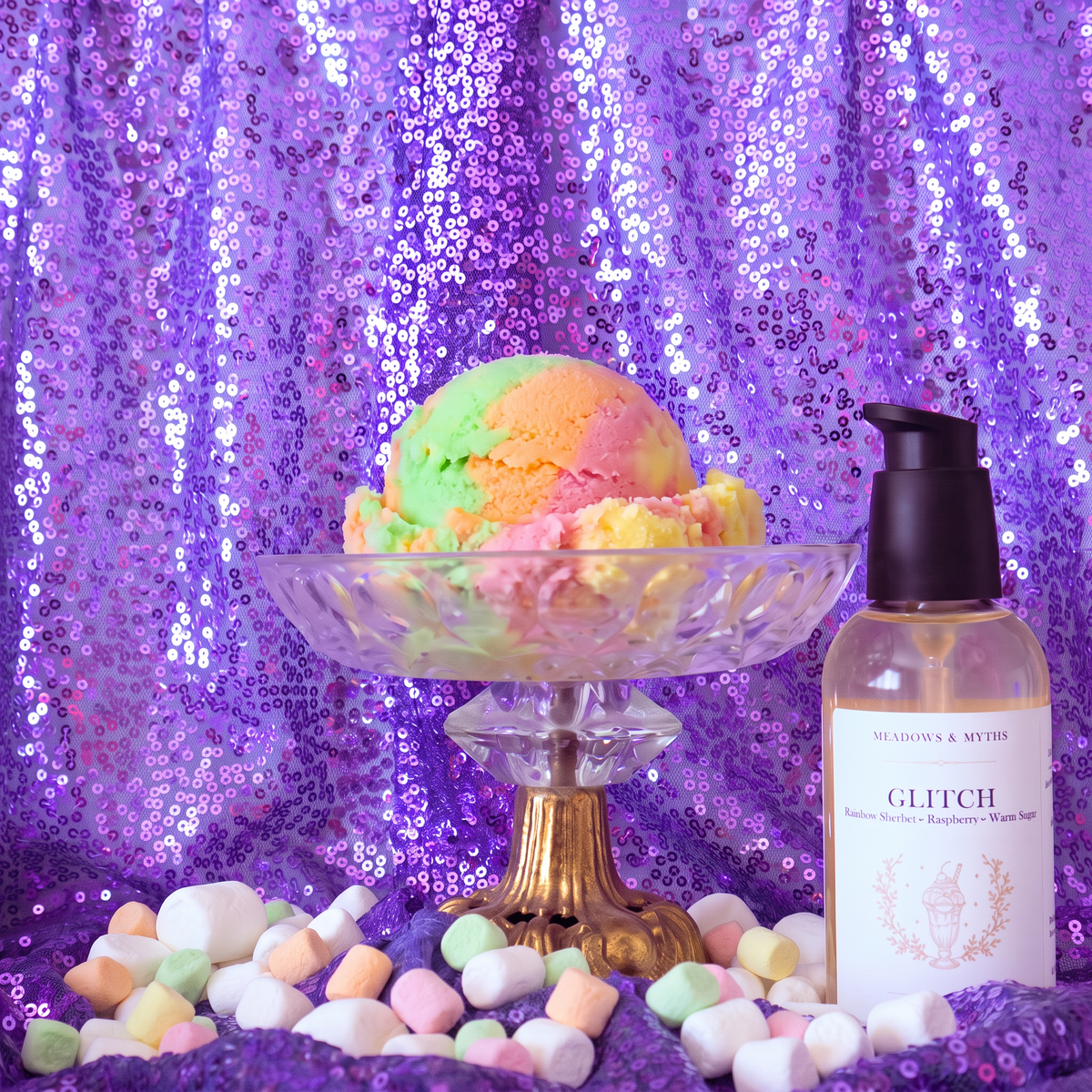 Meadows & Myths 'Glitch' body oil bottle. Sitting on purple sequins fabric with the same fabric draped in the background. The glass raised dish has a scoop of rainbow sherbet. Colorful marshmallows are scattered on the ground.  