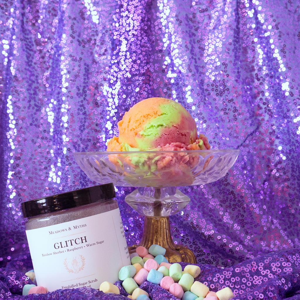 Meadows & Myths 'Glitch' jar of sugar scrub. Sitting on purple sequins fabric with the same fabric draped in the background. The glass raised dish has a scoop of rainbow sherbet. Colorful marshmallows are scattered on the ground.  